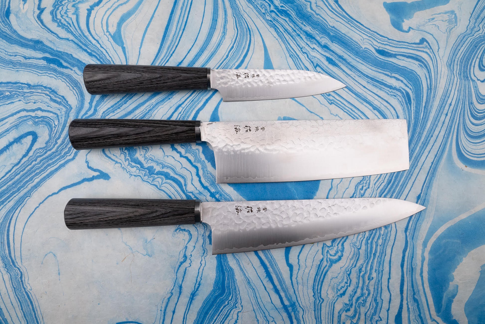Touroku Sakai Gekka 3 Piece Knife Set on a marbled surface, featuring a hand-hammered petty knife, nakiri, and gyuto with dark wood handles, showcasing precision craftsmanship and exceptional balance.