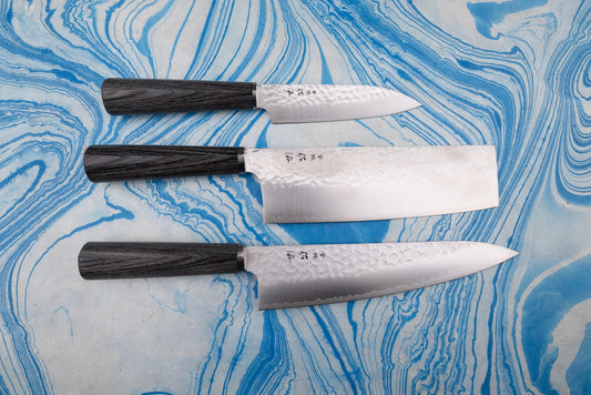 Touroku Sakai Gekka 3 Piece Knife Set on a marbled surface, featuring a hand-hammered petty knife, nakiri, and gyuto with dark wood handles, showcasing precision craftsmanship and exceptional balance.